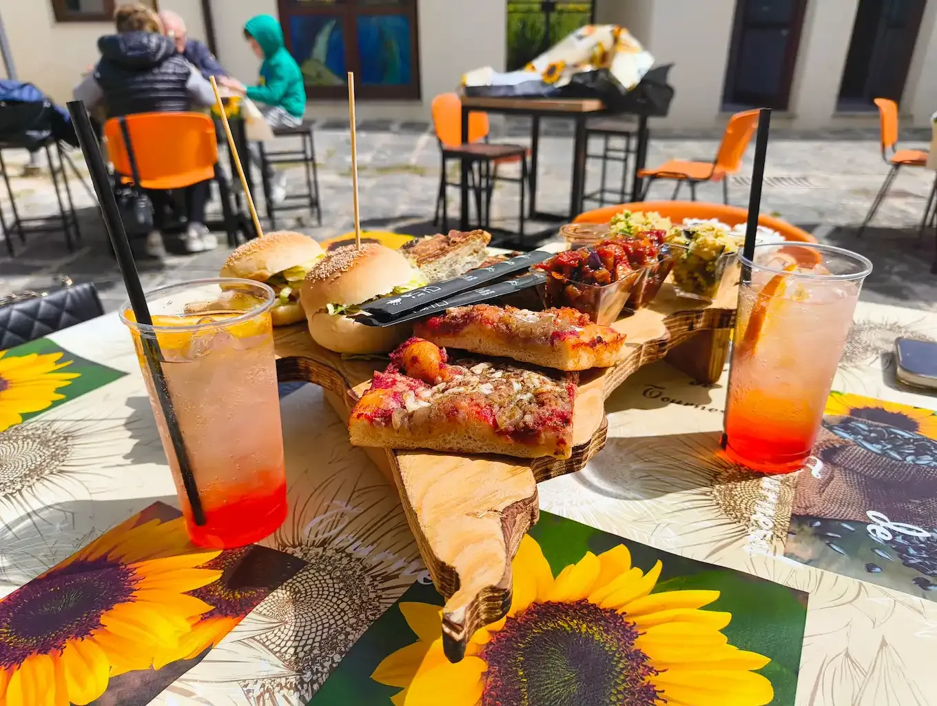 Un delizioso aperitivo all'aperto con una degustazione di piatti tipici, tra cui cous cous con verdure al curry, caponatina di melanzane, tortillas di patate e cipolle, sandwich con prosciutto cotto e maionese al basilico, sushi e sfincionello alla siciliana. Il tutto accompagnato da uno Spritz rinfrescante e acqua